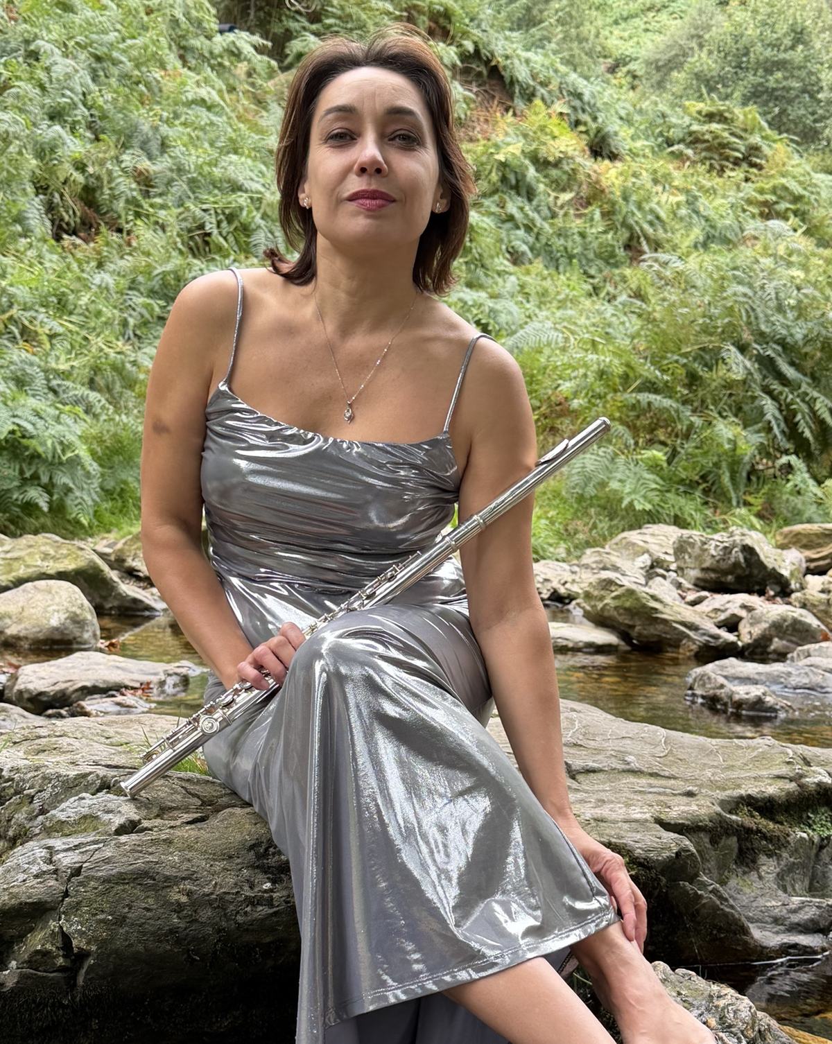 Portrait of Olena Topolskaya, a flautist, sitting on a rock by a mountain stream with her flute, in a silver dress
